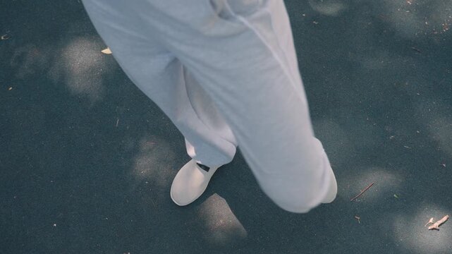 Close up of person lower legs and feet in motion wearing light gray sweatpants and white slip on shoes. scene shows subtle movement against outdoor asphalt surface with scattered leaves