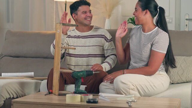 Cheerful young couple high fiving each other in celebration after successfully assembling new flat pack wooden furniture together in their cozy living room apartment setting