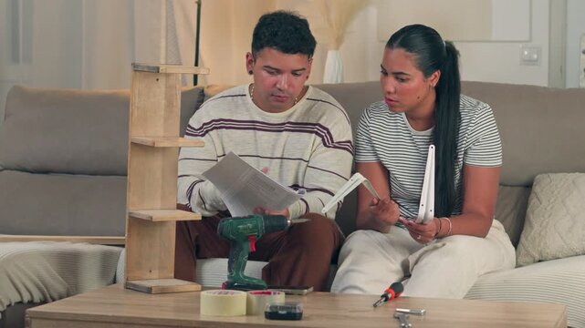 Concentrated young couple sitting on a couch focused on reading a confusing furniture manual booklet together during a domestic home improvement project in a bright apartment setting