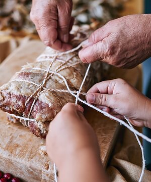 Two pairs of hands tying butcher string around a roast on a scarred wooden board, a tactile intergenerational moment for cooking and recipe campaigns