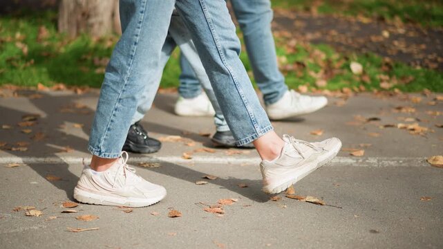 Close up of feet in motion wearing white sneakers and blue jeans on paved path with fallen leaves. Multiple people visible showing different footwear including black shoes. Background