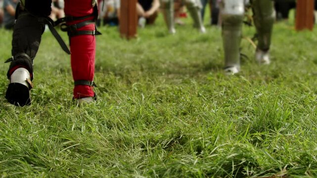 Armored legs move across grass as two knights clash with swords during medieval combat reenactment. Low angle framing emphasizes footwork, plate armor and physical intensity of traditional festival