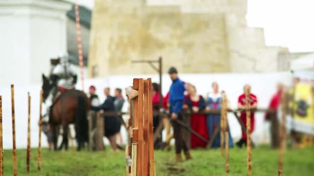 Wooden tournament barriers dominate foreground while armored rider on horseback waits beyond them. Medieval festival crowd and period costumes form lively historical backdrop.