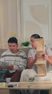 Collaborative young couple sitting on a sofa with the wife handing tools to her husband while he actively works on assembling new wooden furniture in a modern living room interior
