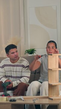 Confident young woman actively constructing flat pack home furniture with tools while sitting beside her partner on a sofa in a modern, stylish apartment living room setup