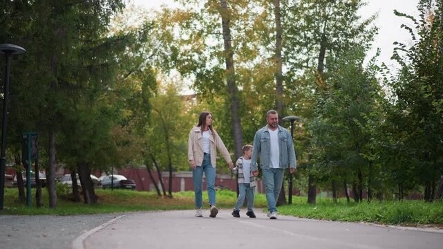 Family of three walking together holding hands in park setting. woman man and young boy walk along paved path surrounded by green trees with some autumn colors visible. Cars are parked