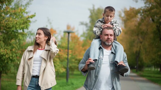 Family walking together in park with man carrying child on his shoulders and woman beside them. Man holds shoes while boy sits securely all three move along path surrounded by green trees