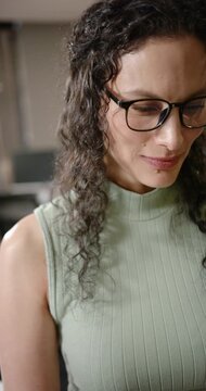 Vertical video: Woman lifting chin, smiling as camera pulling back at desk with green top and specs