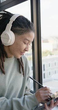 Vertical video: Non-binary African American artist working on tablet with stylus, headset by window