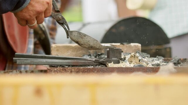 closeup medieval artisan pouring silvery liquid molten metal from casting ladle held with blacksmith tongs into metal mold over charcoal hearth.