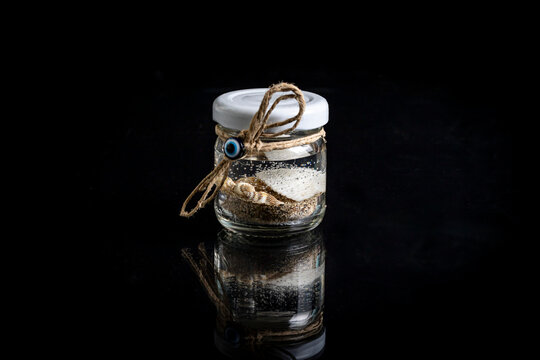 Mini glass souvenir jar with sea sand, seashells and evil eye bead charm tied with jute string isolated on black reflective background