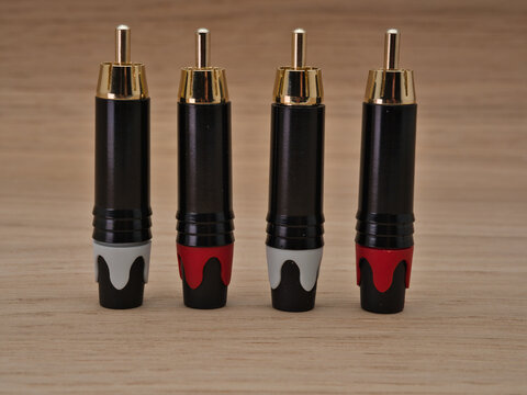 Four RCA Plugs on Wooden Surface - Four audio RCA phono plugs with gold-plated connectors and color-coded boots are neatly arranged on a light brown wooden table.