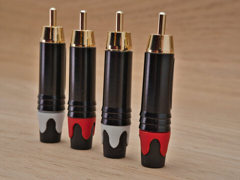 Four RCA Plugs on Wooden Surface - Four audio RCA phono plugs with gold-plated connectors and color-coded boots are neatly arranged on a light brown wooden table.