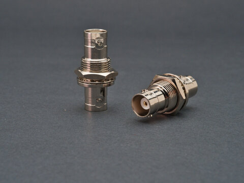 Two Metallic BNC Connectors - Two shiny silver BNC (Bayonet Neill-Concelman) connectors are displayed against a dark gray background, one standing upright and the other l
