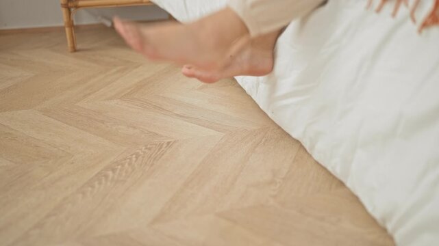 Woman sitting on bed with bare feet placing toes on herringbone wooden floor in a bedroom building; calm morning routine.