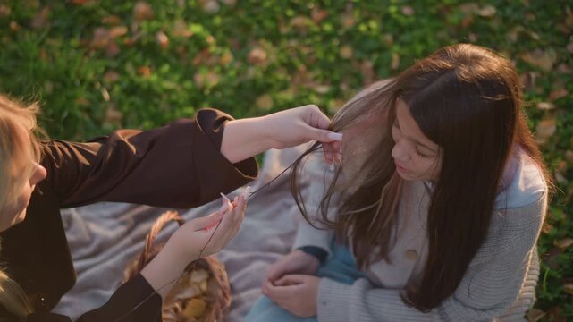 Close up of two women outdoors one holding thin branch with hair wrapped around it while other sits on blanket. Soft natural light illuminates their interaction against green foliage