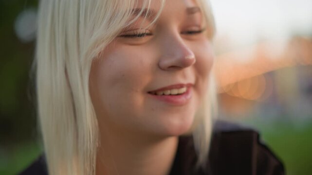 Close up of young woman eating food outdoors with soft natural light. Woman holds and takes bites from item showing long blonde hair and hands with manicured nails. background features