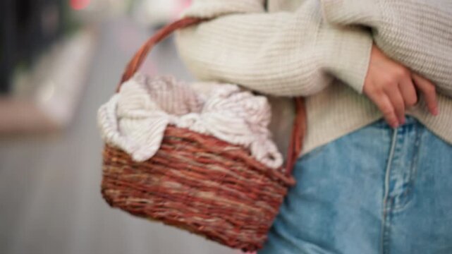 Close up of person wearing blue jeans and holding wicker basket with folded fabric inside walking outdoors. subject hand rests on their hip while camera follows them from behind capturing