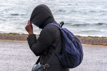 A person wearing a dark hooded jacket and a blue backpack, a smaller worn black bag at the side. Walking along a wet coastal promenade  near choppy a body of water during windy, overcast weather.  © Jacek