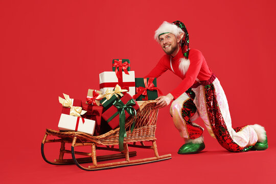 Happy man wearing elf costume pushing sleigh with Christmas gifts against red background