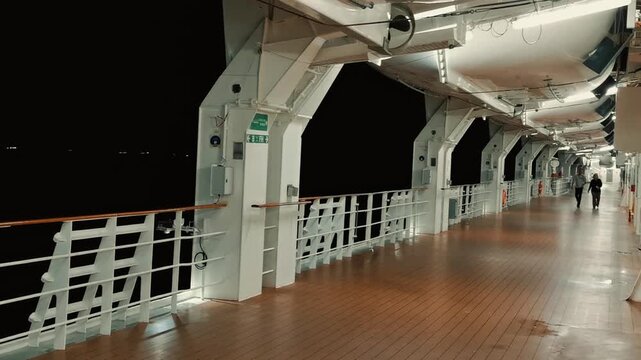 A smooth tilt down reveals an illuminated outdoor promenade deck of a luxury cruise ship at night, with lifeboats mounted overhead and passengers strolling along the wooden walkway.

