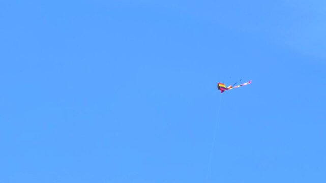 A vibrant, colorful stunt kite with long tails flying and swaying in the wind against a solid, clear blue sky.