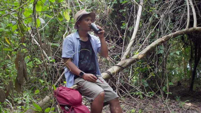 Hiker sitting on a fallen log in a lush forest, drinking water from a bottle while taking a break from trekking, enjoying the peaceful surroundings and refreshing outdoor atmosphere.