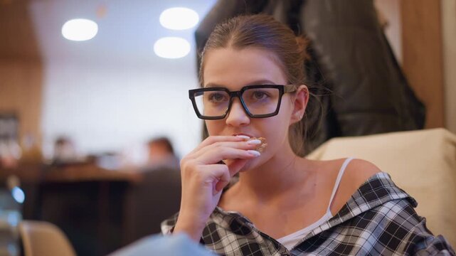 Close up of young woman with glasses eating food in indoor setting. Woman wears casual clothing and holds item near her mouth while soft lighting creates warm ambiance. Background shows