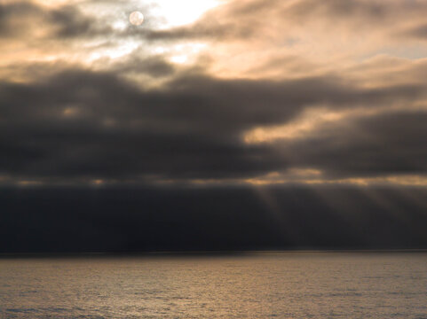 Moody sunset over the ocean with dramatic sunbeams piercing through dark, heavy clouds and illuminating the water along the Oregon Coast.