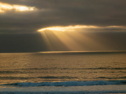 Moody sunset over the ocean with dramatic sunbeams piercing through dark, heavy clouds and illuminating the water along the Oregon Coast.