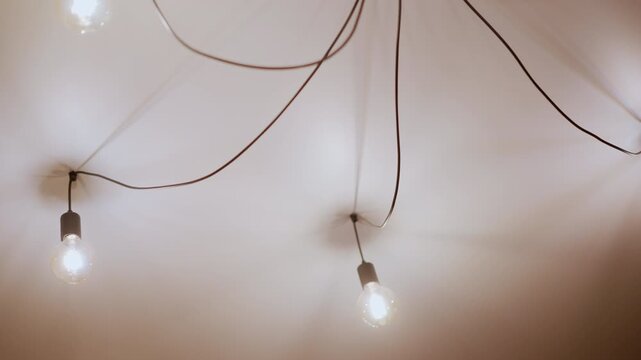 Close up of multiple exposed light bulbs hanging from ceiling with visible wires softly glowing and creating warm ambient lighting. Bulbs are suspended at varying heights against plain white