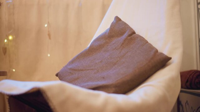 Close up of brown fabric pillow resting on off white chair with soft lighting and string lights in background. scene features warm ambient light creating cozy indoor atmosphere with visible