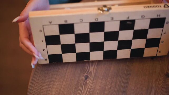 Close up of hands opening wooden chess set box revealing scattered pieces and plastic bag then closing lid to display empty black white checkered board on table