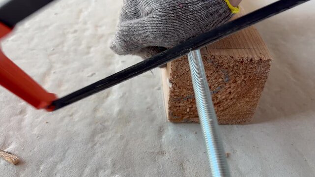 Worker's hands cuts a threaded metal rod with a hacksaw on a block of wood. Manual labor, metalworking, DIY projects