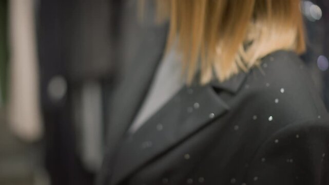 Close up of woman wearing black blazer with small reflective embellishments showing texture and details of her attire. subject stands in indoor setting that appears to be clothing store