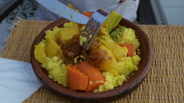 Marrakech, morocco. August 9, 2026. Close up view of a person using a fork and knife to eat a delicious traditional moroccan couscous with meat and vegetables served in a clay pot