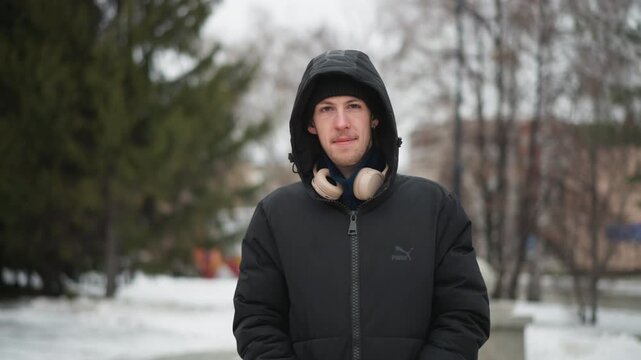 Close up of young man wearing headphones around his neck standing outdoors in winter with snow on ground. Man is dressed in black puffer jacket and hood maintaining stillness throughout