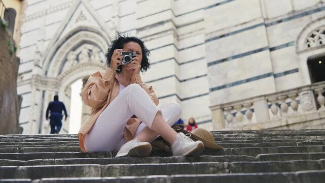 Stylish tourist woman sits on the steps of a historic cathedral in Siena, capturing photos with her retro camera