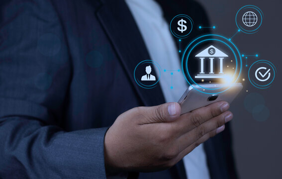 Online mobile banking and global money transfer concept with businessman hand holding smartphone displaying glowing bank icon and connected network nodes on dark background.