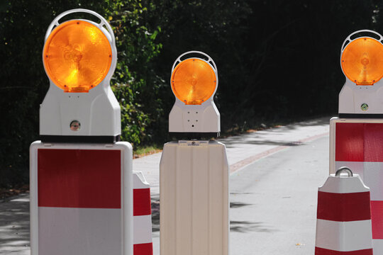 Portable roadwork warning lamps and red white barriers block a narrow street during maintenance. Bright amber lights mark a temporary traffic restriction and construction safety background.