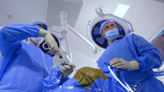Doctors stand still holding the tools and devices at the head of the operated patient. Medics are focused at the screen in front of them. Low angle view at the professionals performing neurosurgery.