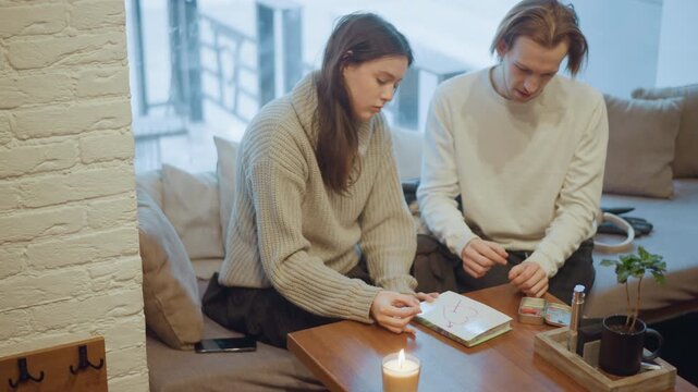 Close up of two people sitting on couch in indoor setting one drawing with colored pencils while other observes. lit candle and small objects like notebooks are visible on wooden table