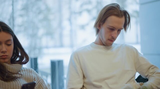 Close up of two young adults sitting side by side indoors each focused on their smartphones. woman wears knitted sweater and man in plain white clothing both maintain steady positions