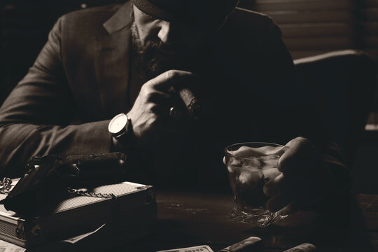 Mafia. Man with cigar, whiskey, gun and case of dollar banknotes at table in dark room, selective focus