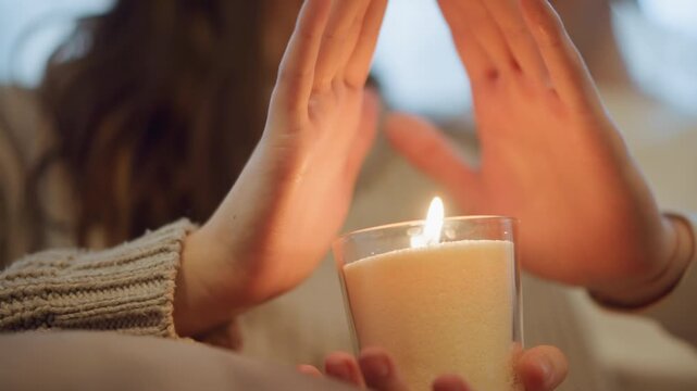 Close up of woman holding lit candle with both hands creating soft warm light. woman wears knitted sweater and gently positions their palms around flame in indoor setting during calm