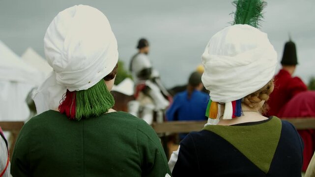 Medium shot captured from behind shows two medieval noblewomen wearing white draped wimples and period dresses watching mounted knight in plate armor during tournament.