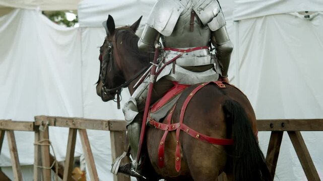 Medium rear view shot shows medieval knight in steel plate armor sitting on brown warhorse with decorative red leather harness by wooden tournament barrier near white camp tents...