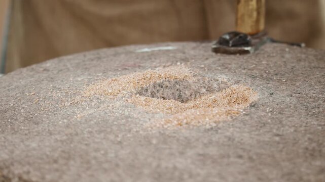 Closeup macro of the central feeding eye hopper of a rotating granite quern millstone filled with whole grain wheat while an artisan's hand grips the wooden handle to grind coarse wholemeal flour...