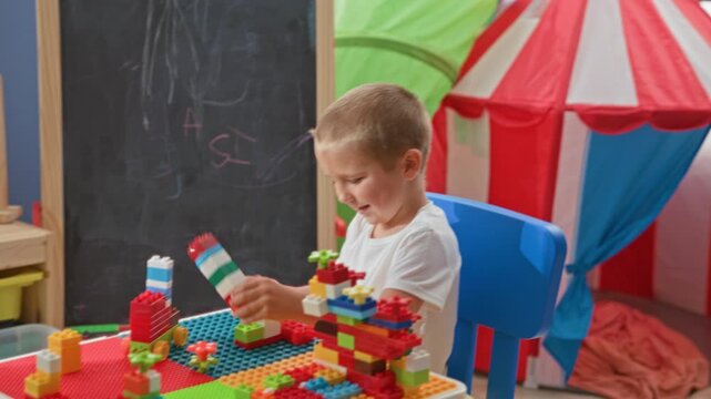 Boy stacking colorful blocks with hand in building playroom, smiling and focused while building a tall tower and holding a wheeled block; joy imagination discovery.