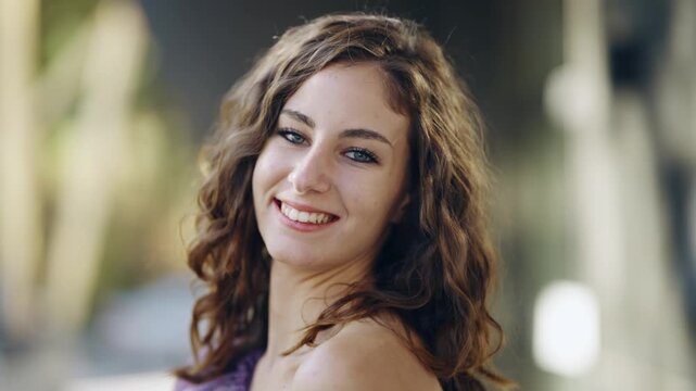 Smiling shy young woman with wavy hair poses outdoors looking at camera over shoulder